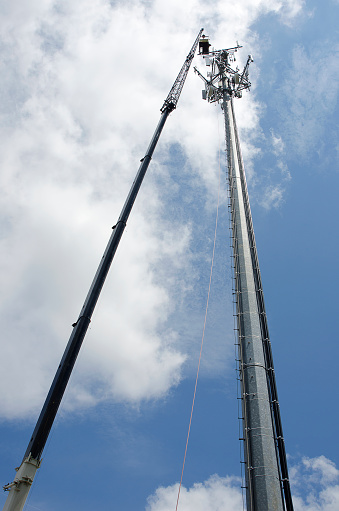 A Cellular/Television/Radio/Communications Tower is being constructed with the aid of a large crane. Workers hang from safety belts and cages over ten stories high to hang communications components to the tower. Presently, there are over 150,000 communications towers constructed in the United States.