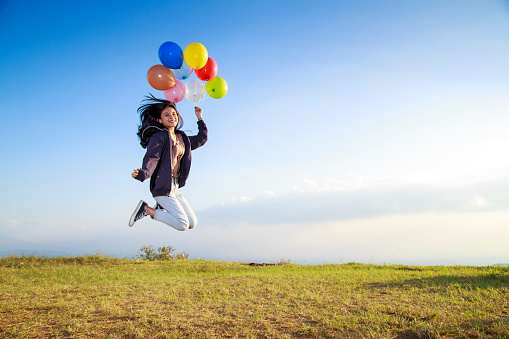 Happy young woman holding colorful balloons and flying over a green meadow