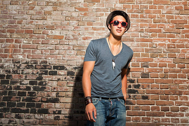 Young man wearing sunglasses near red brick wall Urban asian man with red sunglasses. Good looking. Cool guy. Wearing grey shirt and hat and jeans. Standing in front of brick wall. fashion model men male sunglasses stock pictures, royalty-free photos & images