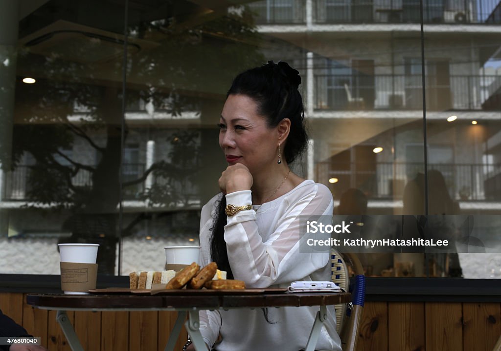 Well-Dressed Japanese Senior Listening at a Tokyo Cafe, Spring Afternoon Well-Dressed Japanese Senior Listening at a Tokyo Cafe, Spring Afternoon. A retired lady with long, black hair and wearing a white blouse and gold bracelet. Enjoying lunch at an outdoor table.  2015 Stock Photo