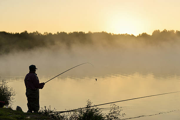 Silhouette of fisherman standing in a lake and catching the fish during foggy sunrise
