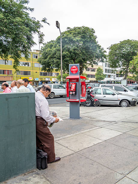 Lima, Peru - January 21, 2015: Male mature Peruvian reading newspaper on Lima street opposite a Claro public payphone. Other people are pictured in the background