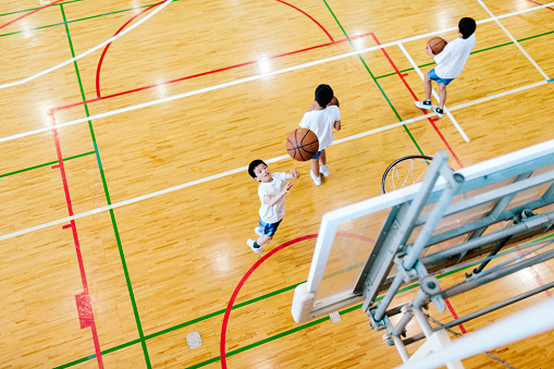 A view of a Japanese high school. A school sports hall with court markings. Children practice their basketball skills with one child shooting directly into the shot. Interior,  horizontal composition. Light reflected on the highly polished floor. 