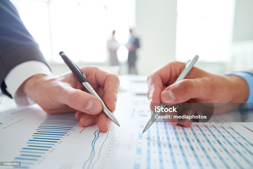 Analyzing statistics Two hands pointing at document with statistical data, defocused business people standing in the background 2015 Stock Photo