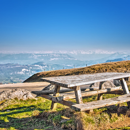 Square composition photography of old picnic wooden table with bench sitting under sunset twilight sunlight, placed in grass and rocks in rolling landscape hill top, with Mont Blanc mountain range massif with snow in background. Scenics landscape view, from top mountain peak, of Alps chain in summer season by clear blue sky. Photography taken on Grand Colombier mountain (near Culoz city, border of Haute-Savoie), in Bugey, in Ain, Rhone-alpes region in France (Europe).