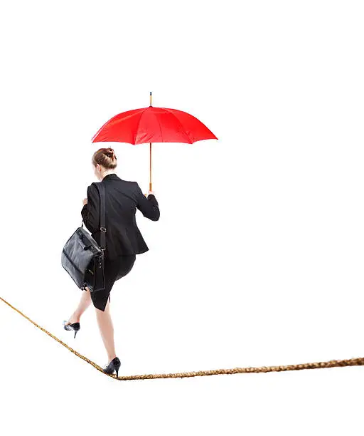 A Caucasian business woman carefully walking on a tightrope with a red umbrella. Business woman balancing work life and home life, navigating her career and future on a dangerous, risky and difficult path. She is walking away from the camera into the future. Photographed in studio on a white background.
