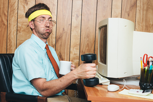 A man and office in 1980's - 1990's style, complete with vintage computer and technology of the time, lifts some small hand weights, getting a short exercise break while remaining at his desk.  He wears a sweatband on his head, taking a short breather from his weight training and drinking some coffee.  Wood paneling on the wall in the background.  Horizontal image.