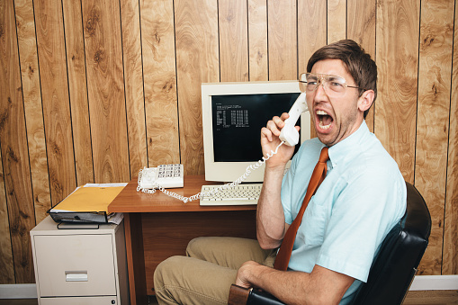 A man and office in 1980's - 1990's style, complete with vintage computer and technology of the time, yells in anger and frustration on the phone to tech support trying to figure out why his computer is broken.  Wood paneling on the wall in the background.  Horizontal with copy space.