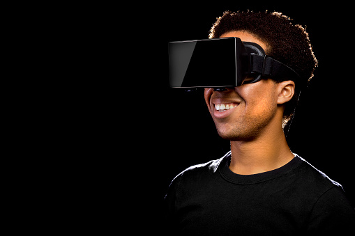 Stock photo of a Virtual Reality headset on a black male playing video games.  The african american male is immersed in a virtual reality experience and on a black background.  He is playing VR games or watcing 3d stereoscopic movies. This image depicts the electronics and video game industry.