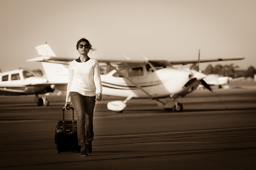 A young woman walks across the tarmac pulling her suitcase. Edited to create and old world travel scene in contrast with the modern aircraft, suitcase, and stylish young woman.