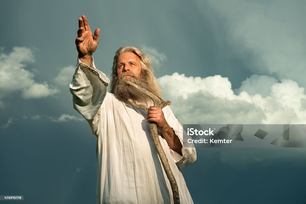 long-haired prophet gesturing in front of dramatic sky An old man with long grey hair and a long grey beard standing in front of a dramatic dark sky. He is wearing a white toga and holding a wooden bar. His arm is stretched out for giving a sign or for blessing and he is looking up to the sky.  God Stock Photo