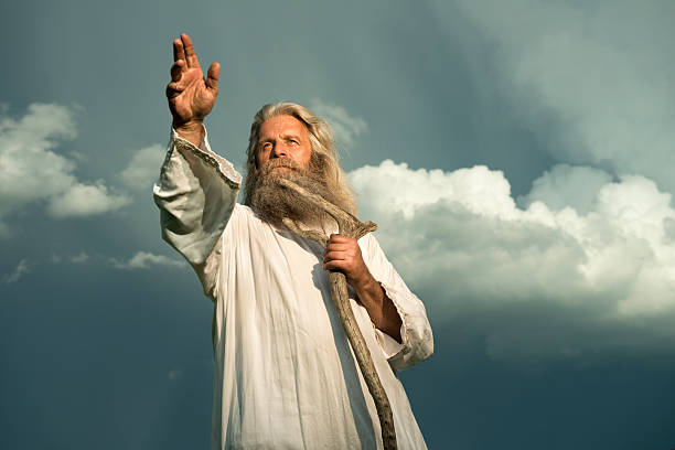 long-haired prophet gesturing in front of dramatic sky An old man with long grey hair and a long grey beard standing in front of a dramatic dark sky. He is wearing a white toga and holding a wooden bar. His arm is stretched out for giving a sign or for blessing and he is looking up to the sky.  apostle worshipper stock pictures, royalty-free photos & images