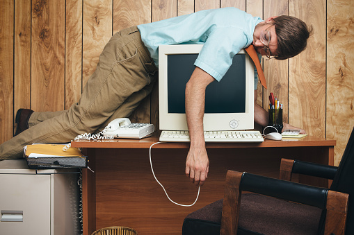 A man and office in 1980's - 1990's style, complete with vintage computer and technology of the time, sleeps on top of his desk, slumping over the computer, too tired and bored to continue working.  Wood paneling on the wall in the background.