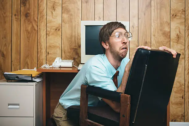 A terrified man and office in 1980's - 1990's style, complete with vintage computer and technology of the time, hides behind his chair at his desk with a look of fear on his face at some crisis or disaster occurring at work.  Wood paneling on the wall in the background.  Horizontal with copy space.