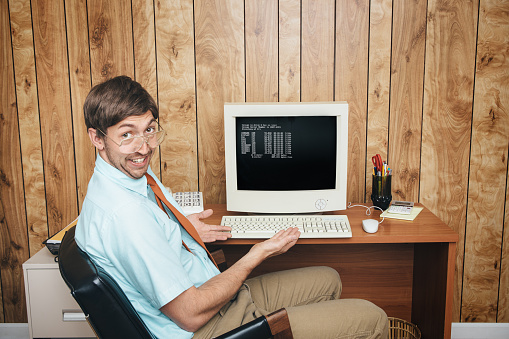 A man and office in 1980's - 1990's style, complete with vintage computer and technology of the time, shows of his state of the art equipment with a look of happiness and pride.  Wood paneling on the wall in the background.  Horizontal with copy space.