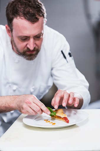 A professional chef in a gourmet restaurant plating food.  He is putting an herb leaf on a dessert plate.  He is wearing a white chef's uniform.