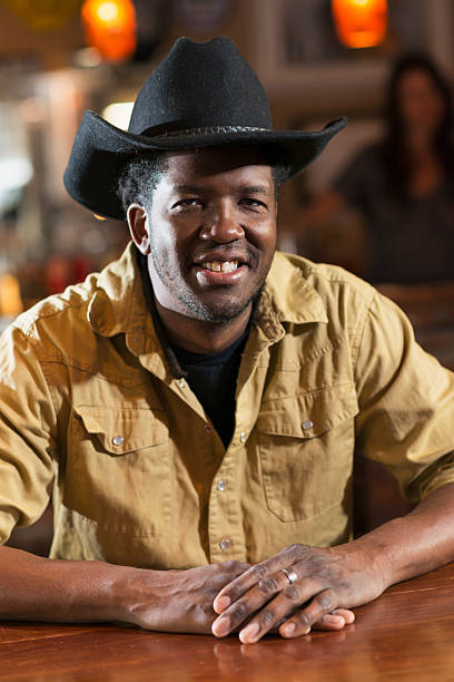 An African American man sitting at a table in a bar restaurant, wearing a black cowboy hat and yellow shirt, smiling at the camera.  The image is a close up of his head and shoulders.