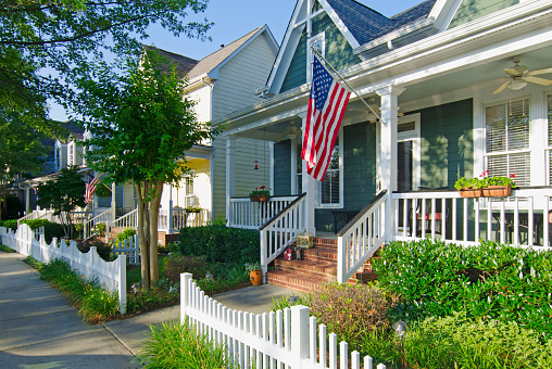 Fort Mill, South Carolina, USA - June 14, 2014: The American Dream is pictured in this iconic image of a row of new, Victorian-style homes with a white picket fence in the Baxter Village neighborhood development located south of Charlotte, North Carolina. Most American homes are now built on smaller lots with sidewalks and tree-lined streets in the suburbs of large cities. An American flag hangs from the front porch in celebration to honor an upcoming holiday. Many Americans express patriotism by flying Old Glory, not only on holidays, but all year long.