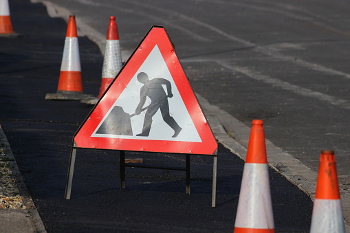 Photo showing a triangle shaped 'Men at Work' road sign, which is sandwiched by rows and rows of red and white bollards.  These traffic cones have been placed on a freshly resurfaced tarmac pavement, so that pedestrians don't walk on it.