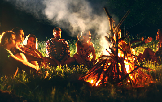 Group of young adults sitting around campfire on summer night. They are at remote place outside the city.There are three guys and four girls,laughing and having fun. The guy on the right is playing guitar.