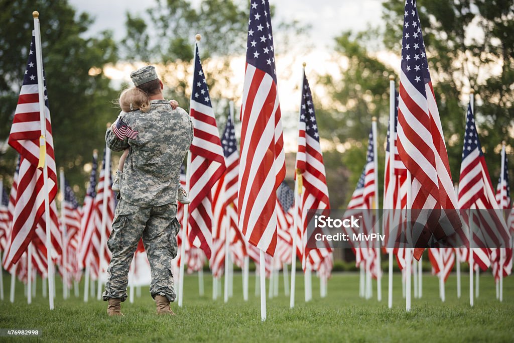 American soldier holding his toddler daughter American soldier who is wearing an American military uniform is holding his daughter in a field of American flags 12-17 Months Stock Photo