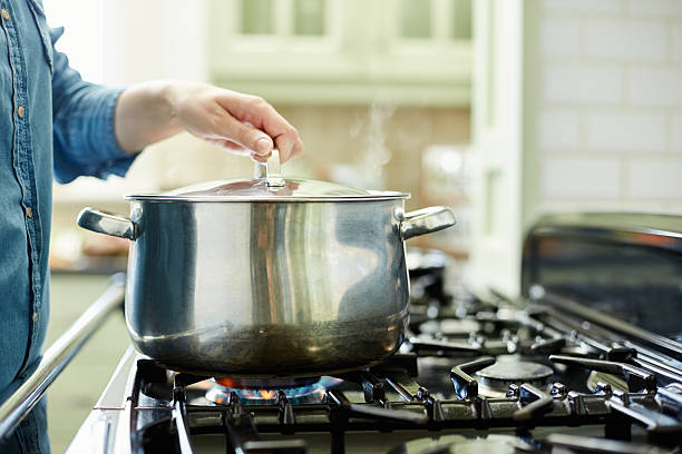 Woman lifting lid on cooking pot in kitchen Cropped image of woman holding lid on cooking pot. Utensil is placed on gas stove. Female is holding stainless steel covering. She is cooking food in domestic kitchen. gas stove burner photos stock pictures, royalty-free photos & images