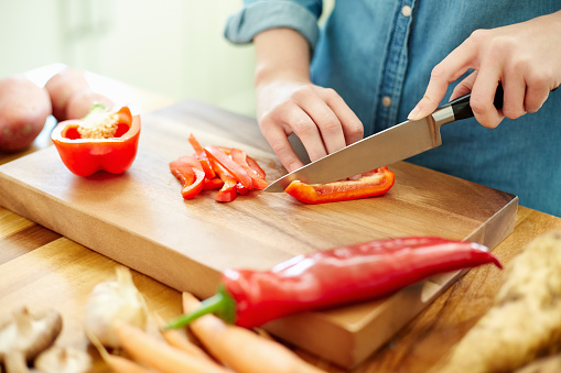 Midsection image of woman cutting red bell pepper on board. Focus is on her hands. Female is preparing food at wooden table. She is in domestic kitchen.