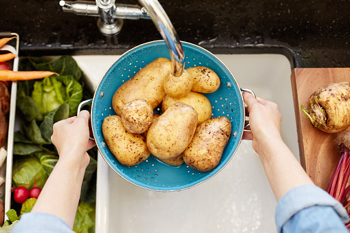 High angle cropped image of hands washing potatoes in colander. There are various vegetables on sink. Woman is holding root vegetable under faucet. She is in domestic kitchen.