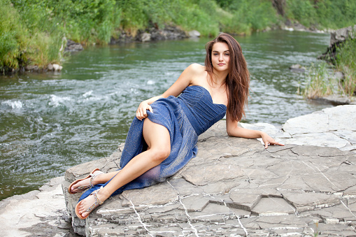 Young attractive brunette in long blue dress is lying on a rock by the river. Blurred background of the river and green nature, she is smiling and looking at the camera. Summer atmosphere. Shooting with Canon EOS 5D Mark II.