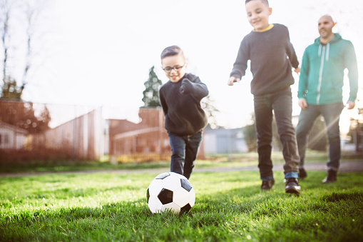 A young Hispanic father smiles and laughs as he and his boys play soccer on a sunny day in the grass of their front yard.  Depiction of a loving dad and family engaging in a healthy lifestyle.  Horizontal with copy space.