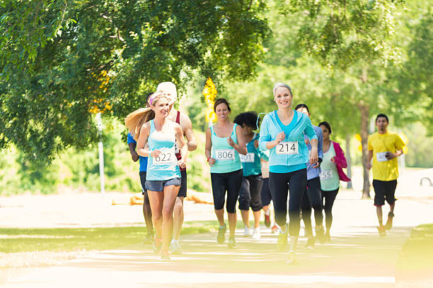 Diverse runners approaching finish line during marathon or 5k race Diverse young adults, mid adults, mature adults, and senior adults are running excitedly toward finish line during marathon or 5k race for charity in sunny park. They are wearing athletic clothing and race contestant numbers. Runners are smiling and running in large group. walking for a cause stock pictures, royalty-free photos & images
