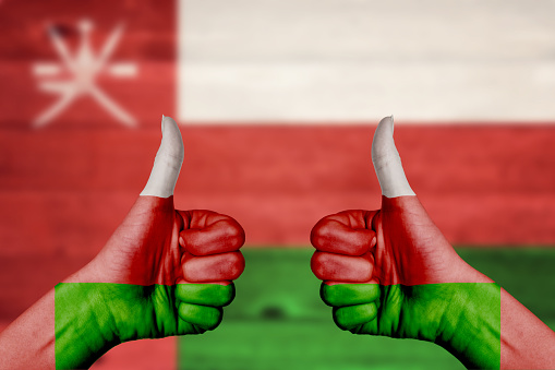 Oman flag painted on female hands thumbs up with blurry wooden background