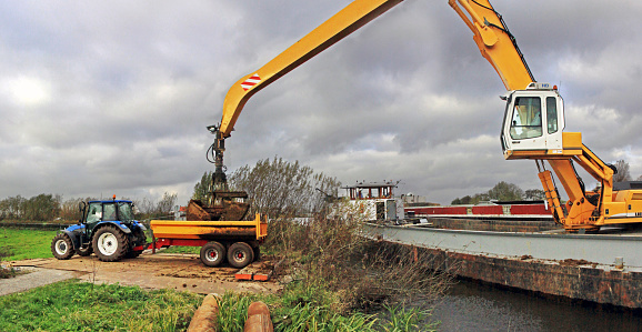 Woubrugge, Netherlands - November 7, 2012: a ship-mounted crane deposits clay from a large barge into a trailer pulled by a tracktor. Part of dike reenforcement work in a polder.  Dark and cloudy sky. 