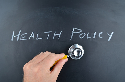 Health Policy handwritten on a school chalkboard in white chalk. A human hand holds a doctors stethoscope nearby.