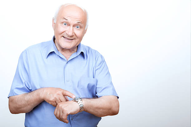 It is high time. Smiling grandfather pointing his wristwatch and looking ahead while standing isolated on white background.