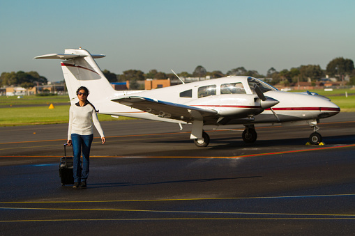 A tourist arrives from her small aircraft. She is walking across the air field with her suitcase in tow. Aircraft visible behind her.