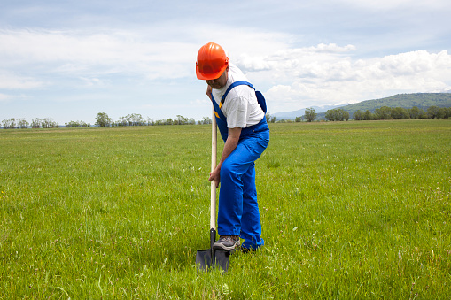 Worker in coveralls is digging with a shovel, in a green meadow. He is starting a new construction project. The image was shooted in a sunny day in the spring. The main object is in the center. There are little clouds on the sky in the background.