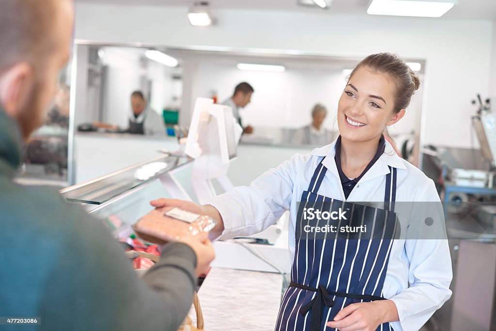working at the butcher's shop a young female trainee butcher tends the meat counter in a butcher's shop. She is wearing a white coat and striped apron and is smiling to a customer across the counter as she hands over a pack of sausages . Butcher's Shop Stock Photo