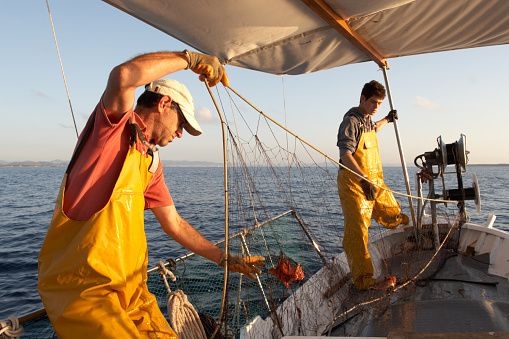 Two fishermen working on a boat in the Mediterranean Sea, in the morning outdoors with the sea in the background. Father and son wearing fishing clothes work together collecting fishing nets.