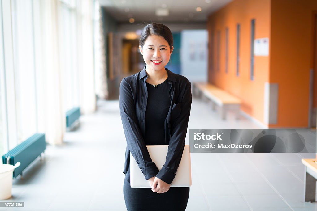 Asian college student standing with a laptop A portrait of an Asian college or university student at campus. She is standing in the middle of the corridor and is holding a laptop. University Student Stock Photo