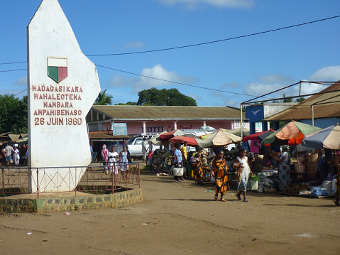 Diego Suarez, Madagascar - May 13, 2015: In the busy town of Mahavanona south of Diego Suarez, a monument celebrating Madagascar's Independence in 1960 stands in the town center as Malagasy men, woman and children walk through the shops and stalls selling fruits, various supplies, clothing and food in the town market.