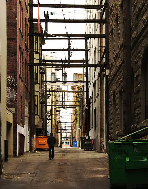 Grungy shot of dumpsters in an urban alleyway with a hooded stranger in the distance.