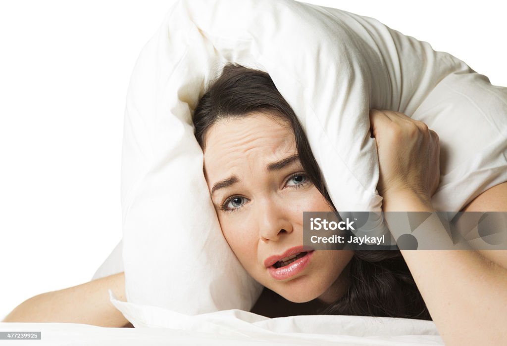 Tired sleepless woman A tired sleepless woman puts the pillow over her head and looks helplessy at camera. Isolated over white. Adult Stock Photo