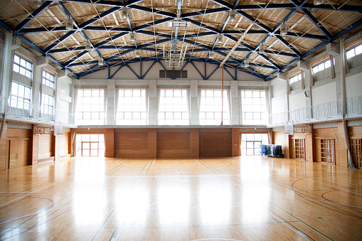 A view of a Japanese high school. An empty sports hall with a vaulted ceiling and court markings. Interior shot, nobody, horizontal composition. Light reflected on the highly polished floor. 