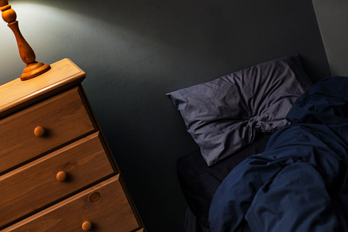 Illuminated incandescent lamp lighting a generic dark evening bedroom from the top of a light brown wooden clothes dresser bureau. The various shades of blue on the sheet, pillow, and duvet blanket look warm and cozy despite being scattered and messy on the unmade twin sized bed.