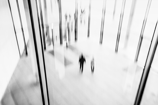 abstract black and white image of two male and female people, couple, walking in modern lobby, in the background can be seen large glass wall and glass doors, elevated view, long exposure, blurred motion