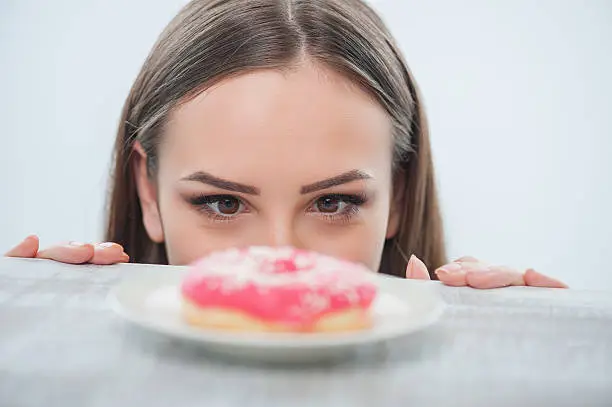 Beautiful girl is looking at unhealthy donut with appetite. It is situated on a table. Isolated on a white background