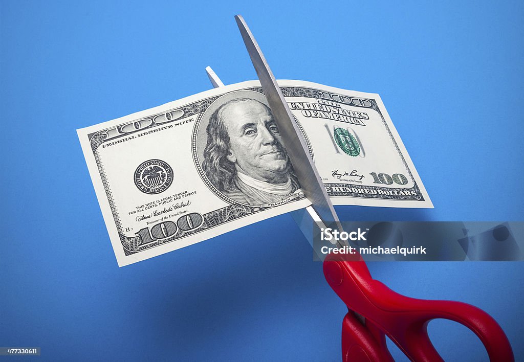 Cutting costs 100 dollar bill being cut with scissors isolated on a blue background Cutting Stock Photo