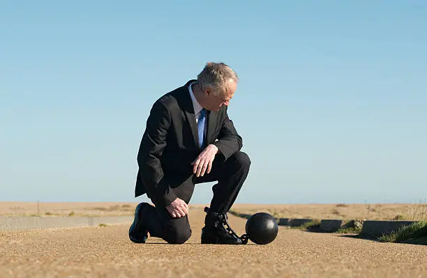 Businessman kneeling down on a remote road, with a ball and chain attached to his ankle. 