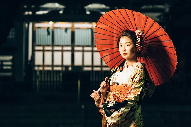 Beautiful japanese woman stand inside of traditional japanese temple. She wears kimono, obi and hold oil paper umbrella.The kimono is very colorful and elegant. She seems very pensive. There is beautiful kanzashi in her hair.